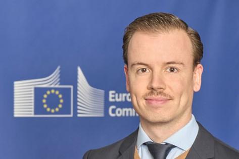 Close-up of a young man with a moustache and short brown hair, with the European Commission logo in the background.